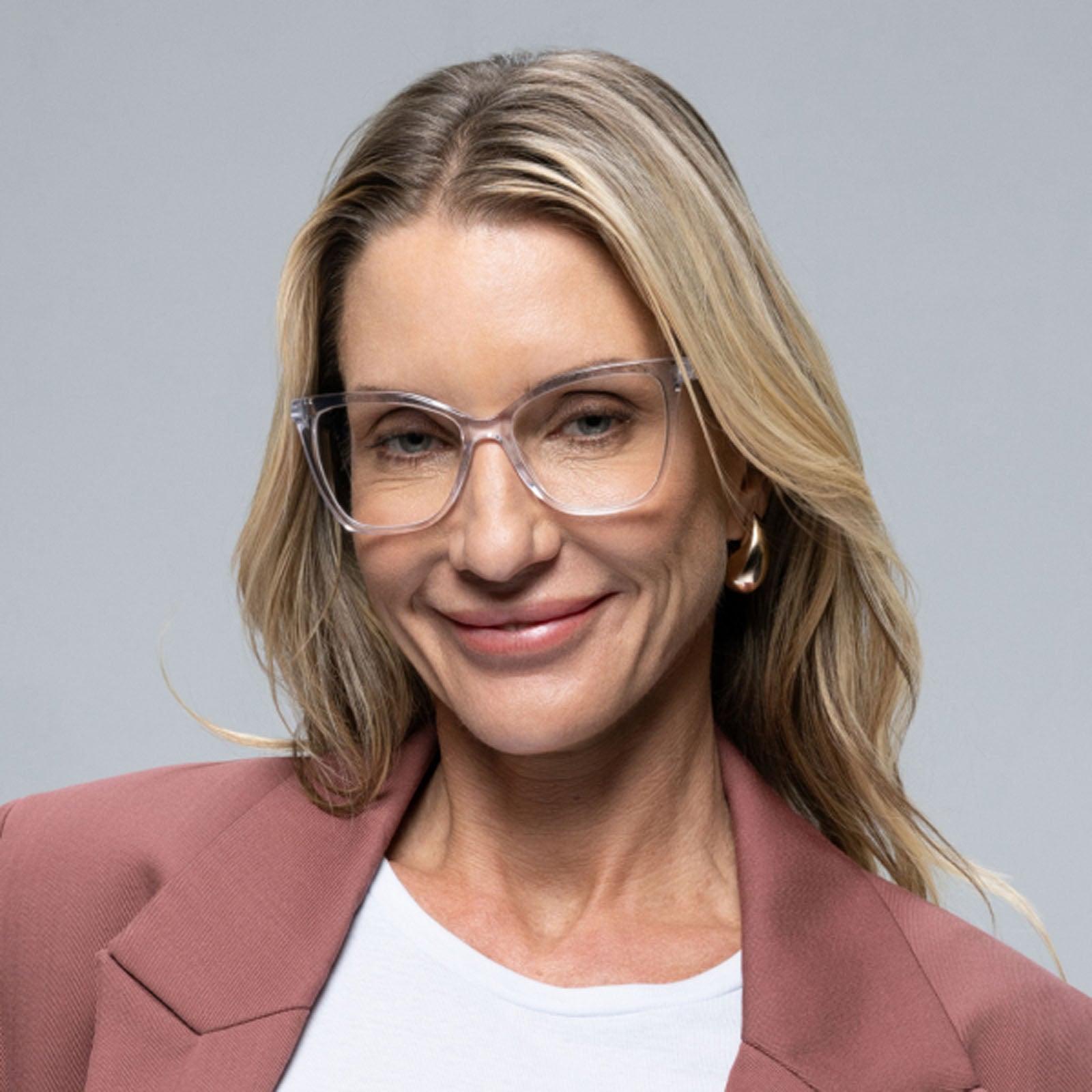 A woman with long blonde hair and clear glasses smiles at the camera in a mauve blazer, modeling Lucyd's Moonrise women's sunglasses with polarized purple gradient lenses against a plain gray background.