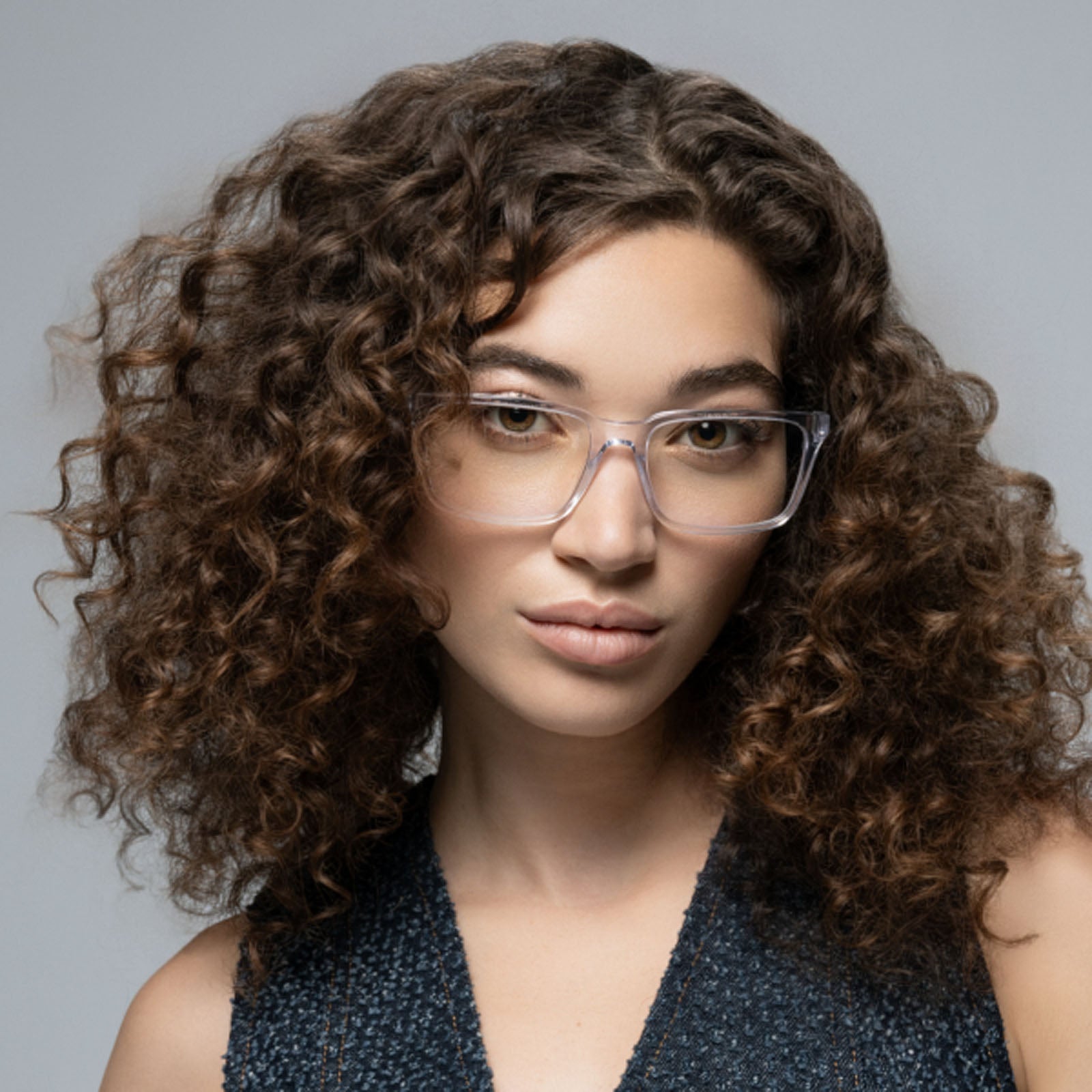 A young woman with voluminous curly brown hair models Lucyd's Eclipse clear-framed smart eyewear, wearing a sleeveless dark textured top against a plain gray background.