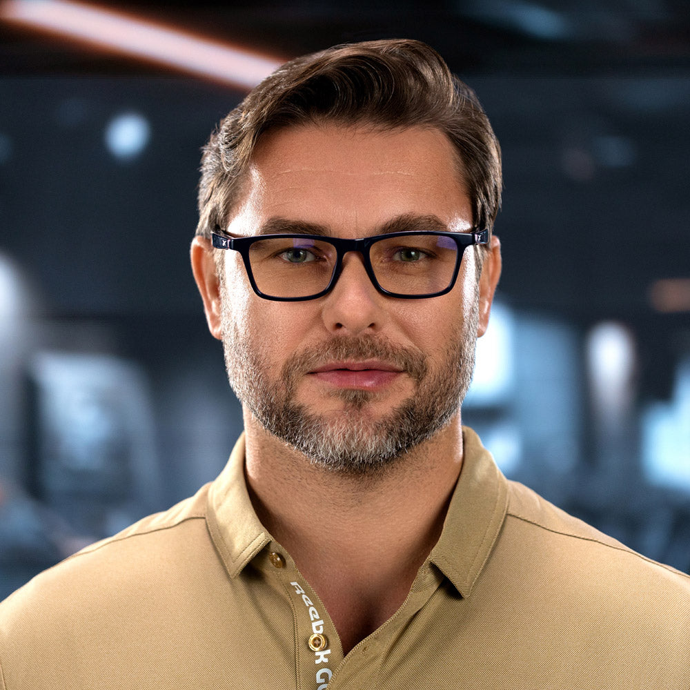 A man with short brown hair, a trimmed beard, and glasses featuring open-ear audio wears the Reebok Dash in front of a softly-lit, blurred indoor background.