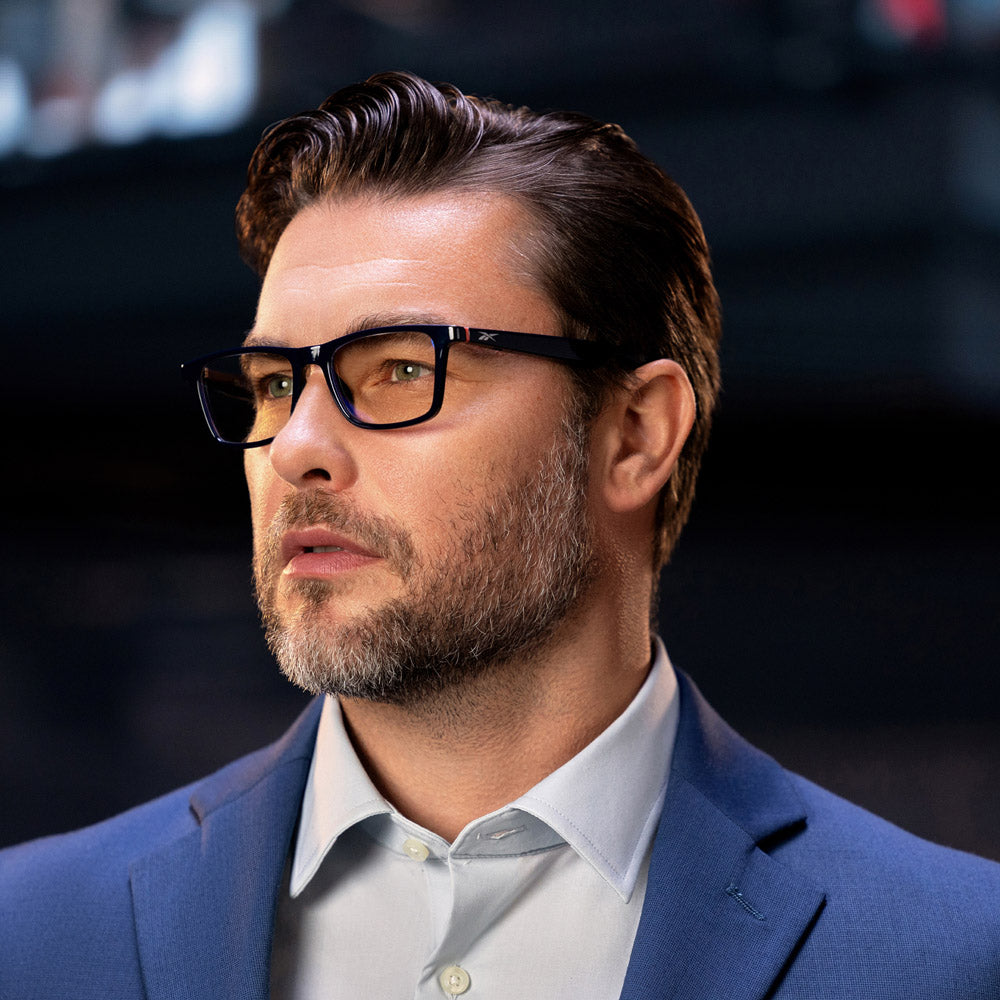 A man with brown hair, a beard, and glasses with blue light protection wears the Reebok Dash blue suit jacket and light blue shirt, gazing off to the side against a dark, blurred background.