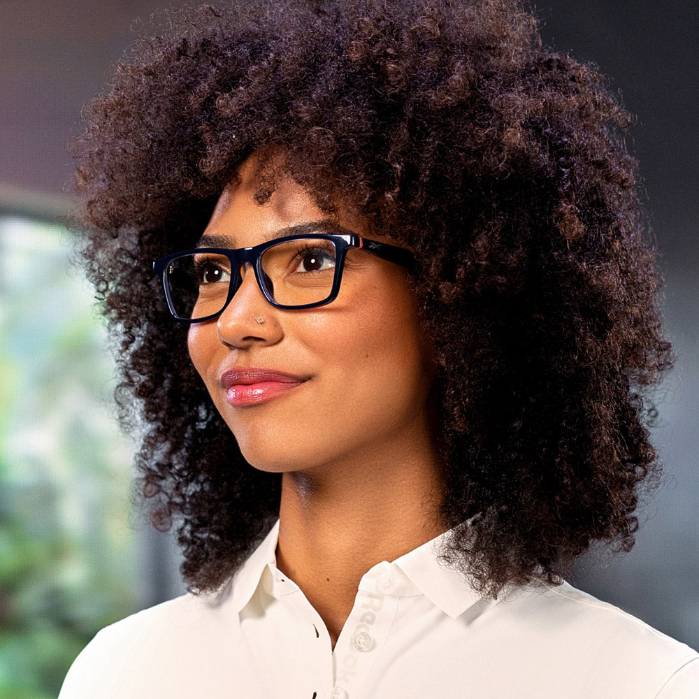 A woman with curly hair wears Reebok Dash smart glasses and a white collared shirt, smiling softly as she looks to the side against a blurred green and gray backdrop.