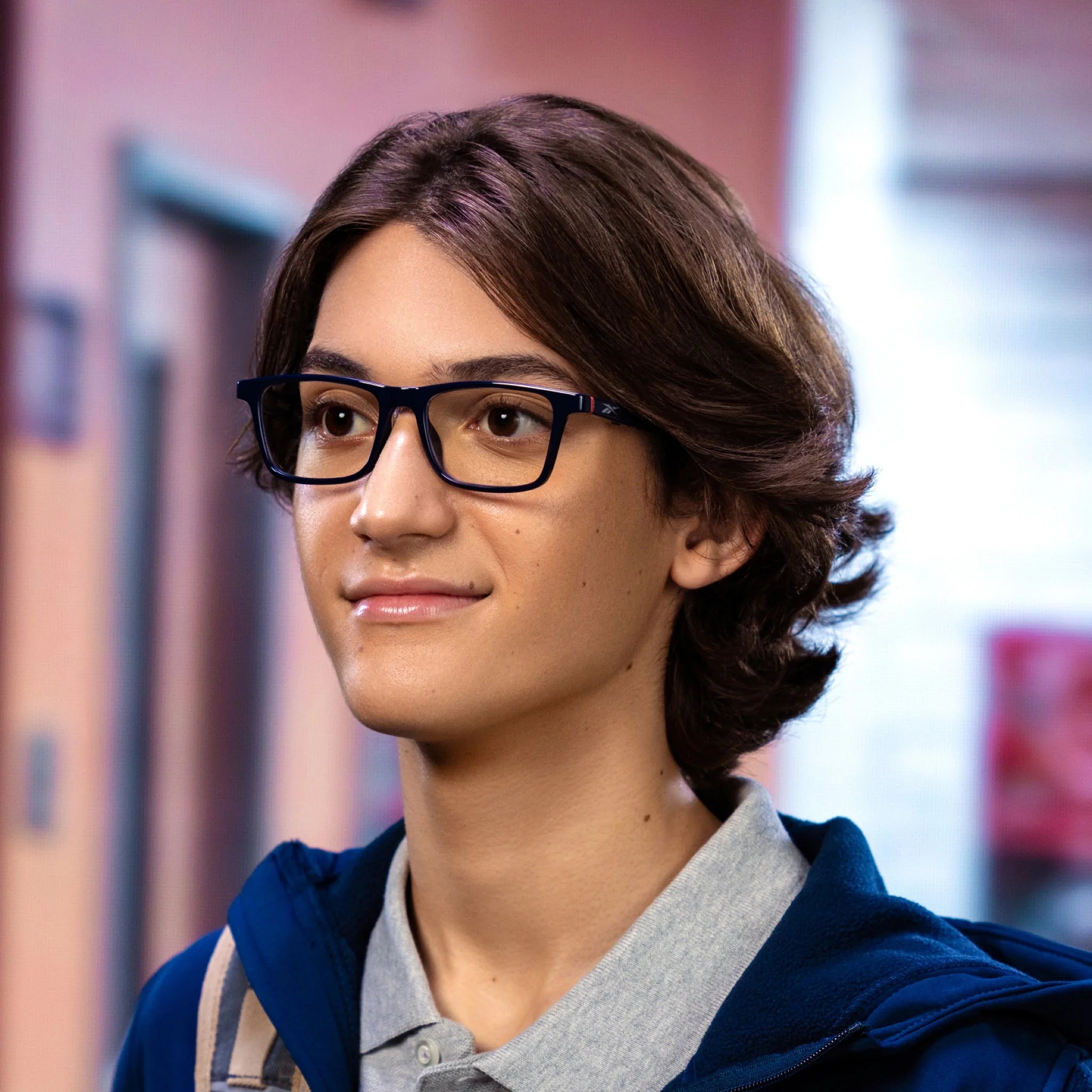 A young person with medium-length brown hair and glasses, wearing a blue jacket and gray collared shirt, stands indoors with a slight smile—perhaps enjoying open-ear audio from their Reebok Dash smart glasses, with a blurred hallway behind them.