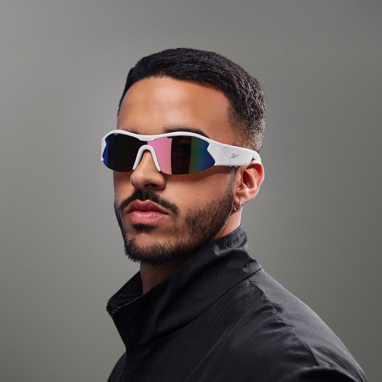 A man with short black hair and a trimmed beard poses against a plain gray background, wearing Reebok Flash white Bluetooth sunglasses with reflective lenses and a black zip-up jacket.