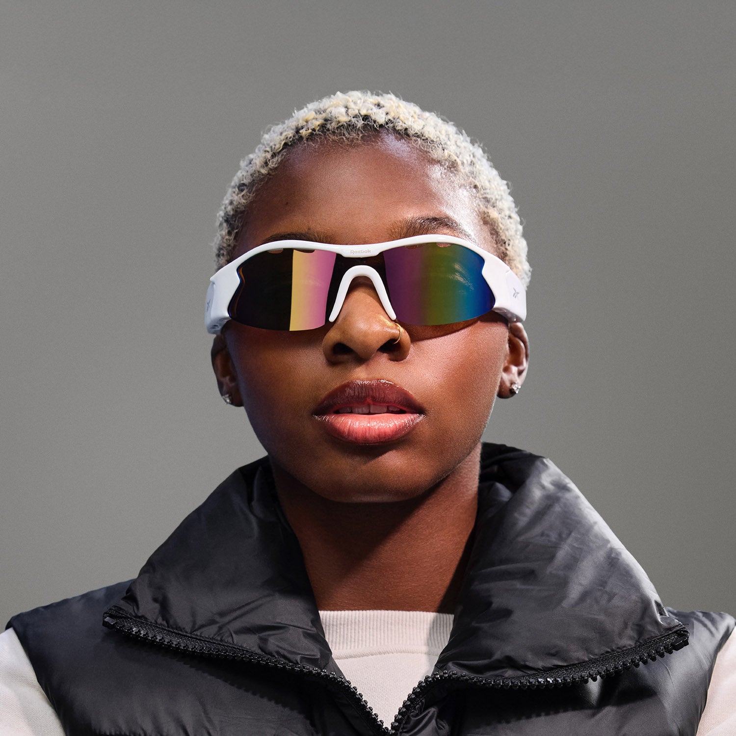 A person with short, bleached blonde hair wears Reebok Flash audio sunglasses with polarized lenses and a black puffer vest over a light shirt, posing against a plain gray background.