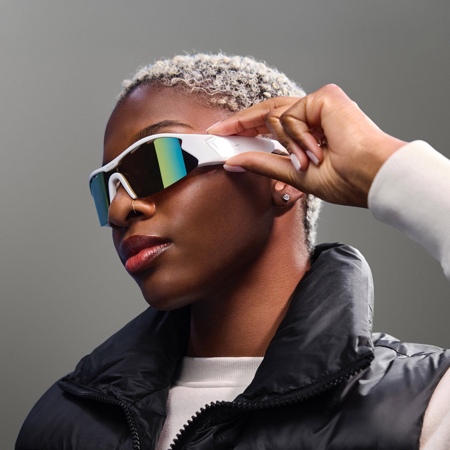 A person with short, light blonde hair wears Reebok Flash Bluetooth sunglasses with mirrored lenses and a black puffer vest, touching the side of the glasses against a plain gray background.