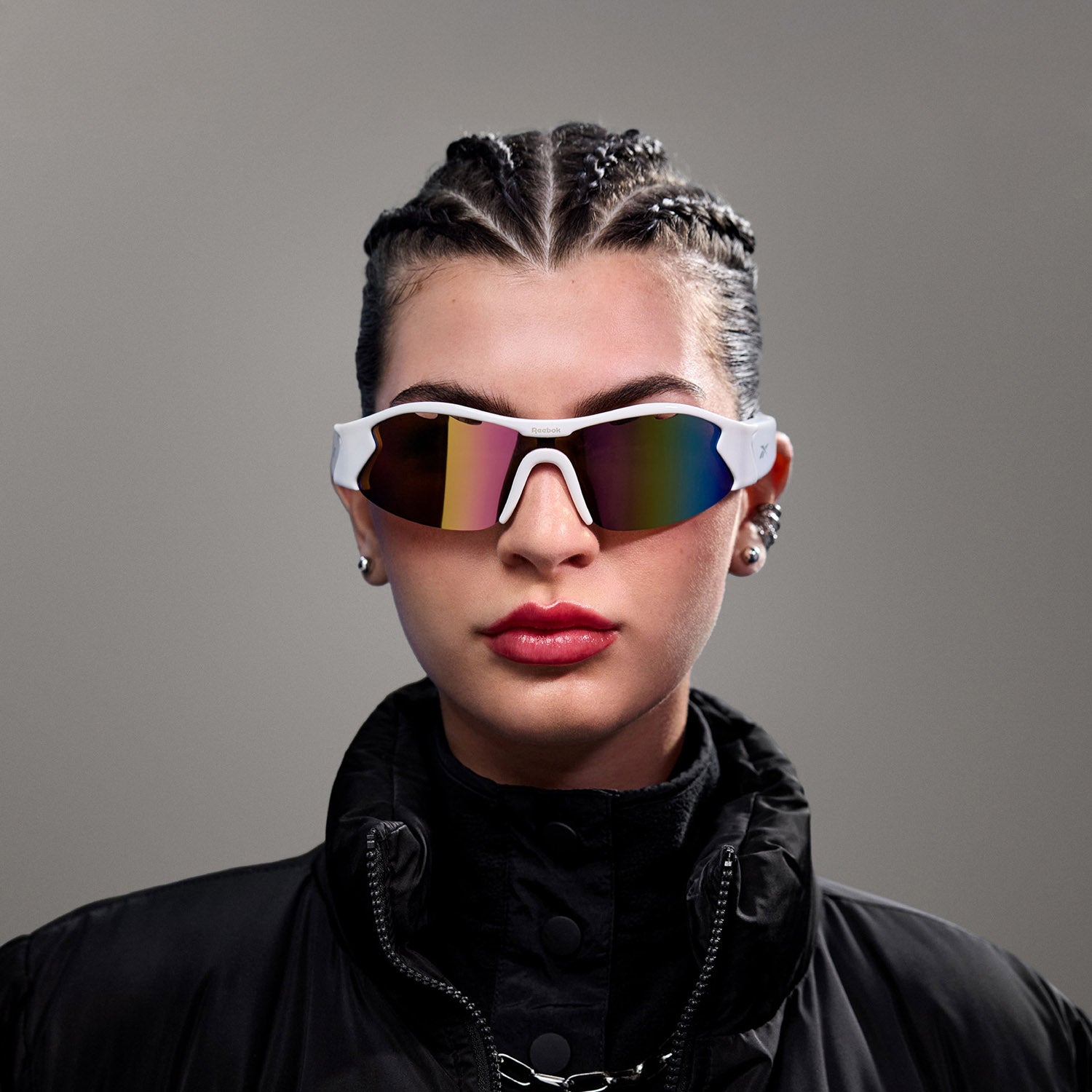 A person with braided hair models Reebok's Flash Bluetooth sunglasses with polarized lenses, red lipstick, earrings, and a black high-collared jacket against a plain gray background.