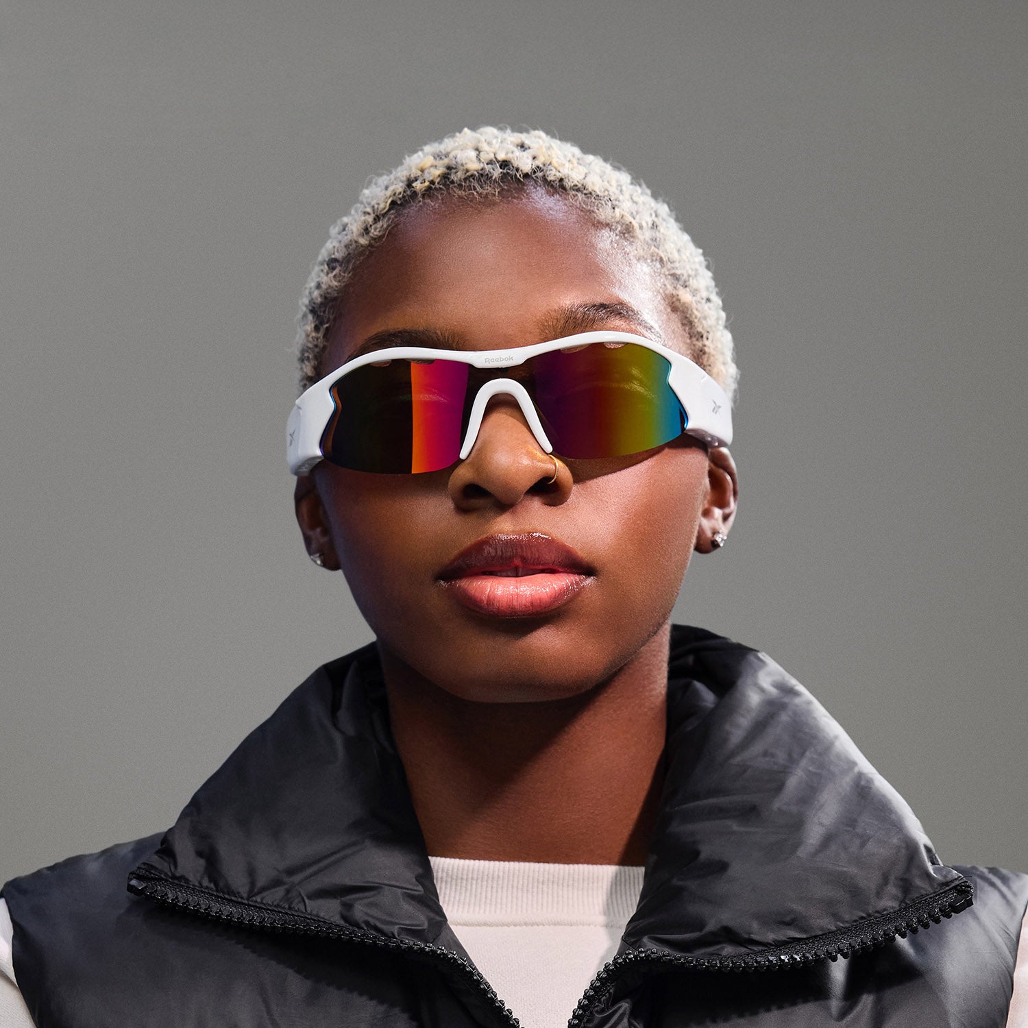 A person with short, bleached hair models Reebok Flash white Bluetooth sunglasses with rainbow-tinted lenses, a black puffer vest, and a light top against a plain gray background.