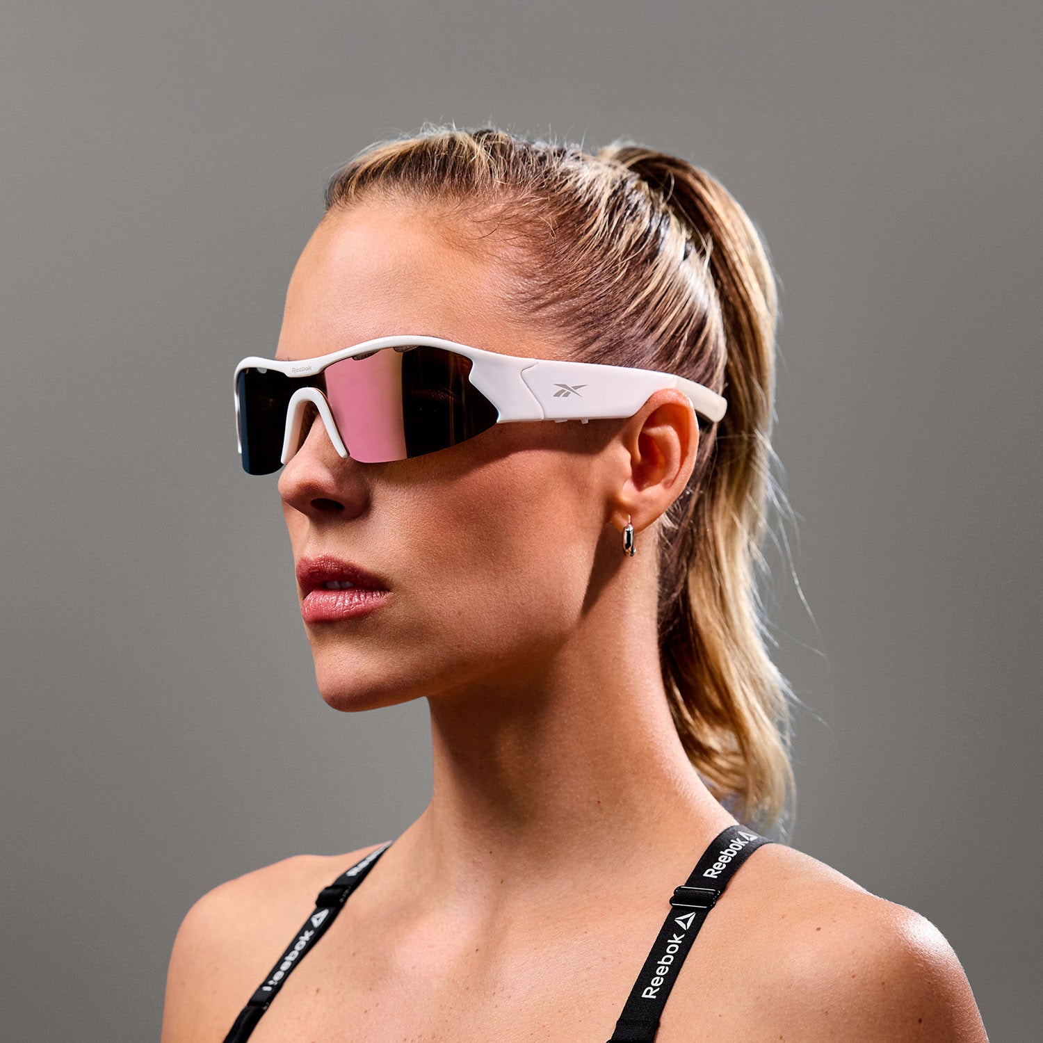 A blond woman with a ponytail wears Reebok Flash white Bluetooth sunglasses with mirrored lenses, a small hoop earring, and a black sports top with Reebok straps, posing against a plain gray background.