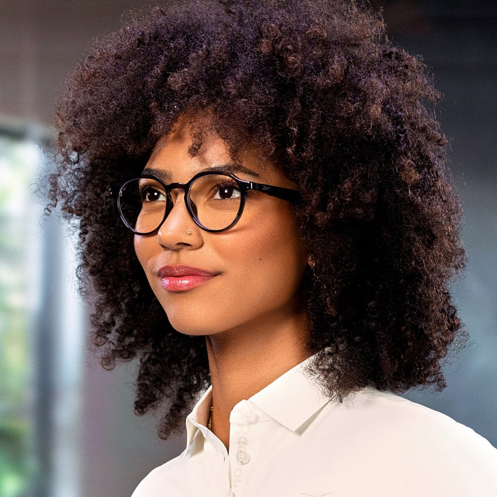 A woman with curly dark hair and round glasses, dressed in a white collared shirt, smiles gently while looking to the side. She wears Jet eyewear by Reebok, which offers privacy features. The softly blurred background provides a calm ambiance.