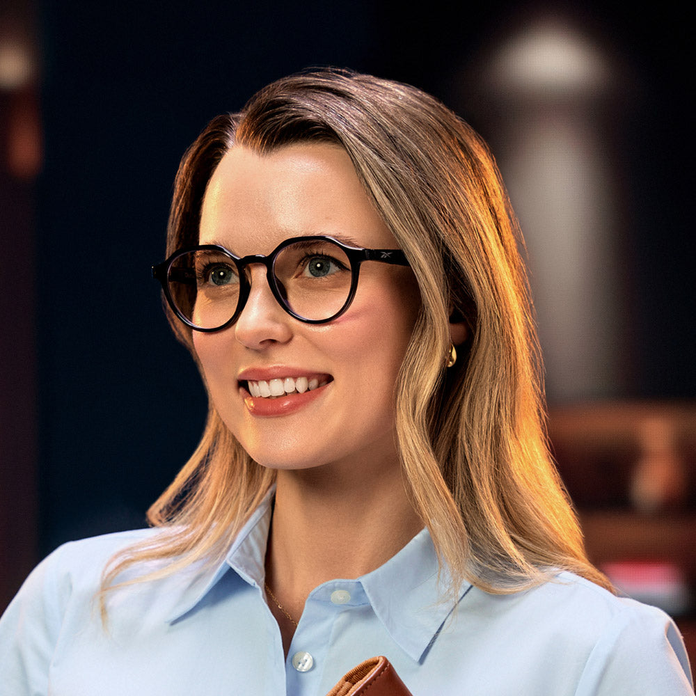 A smiling woman with long blonde hair wears smart glasses and a light blue button-up shirt, holding the Reebok Jet product indoors against a softly blurred background.