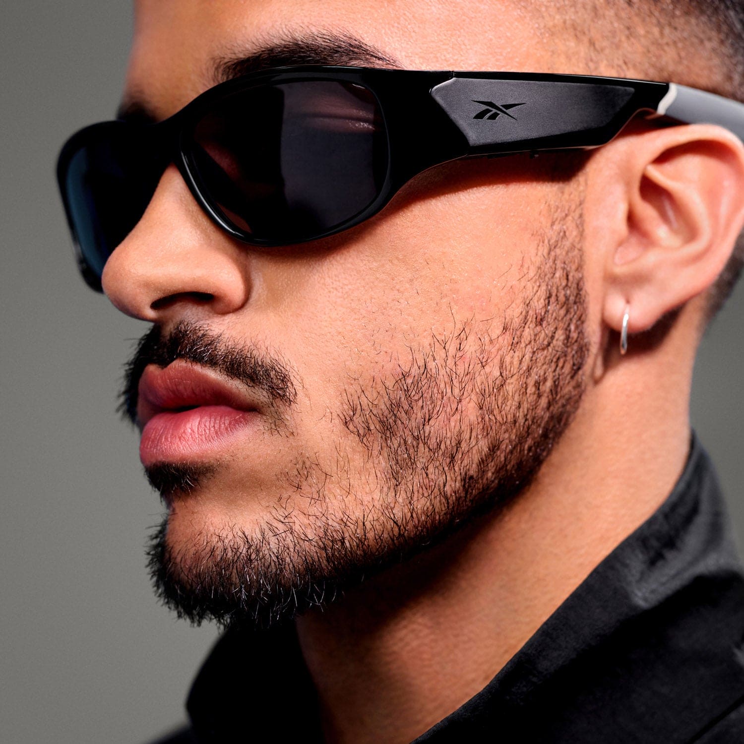 A close-up of a man wearing Reebok NITROUS sunglasses with polarized lenses, a hoop earring, and a black collared shirt. He has a neatly groomed beard and mustache, set against an out-of-focus gray background.