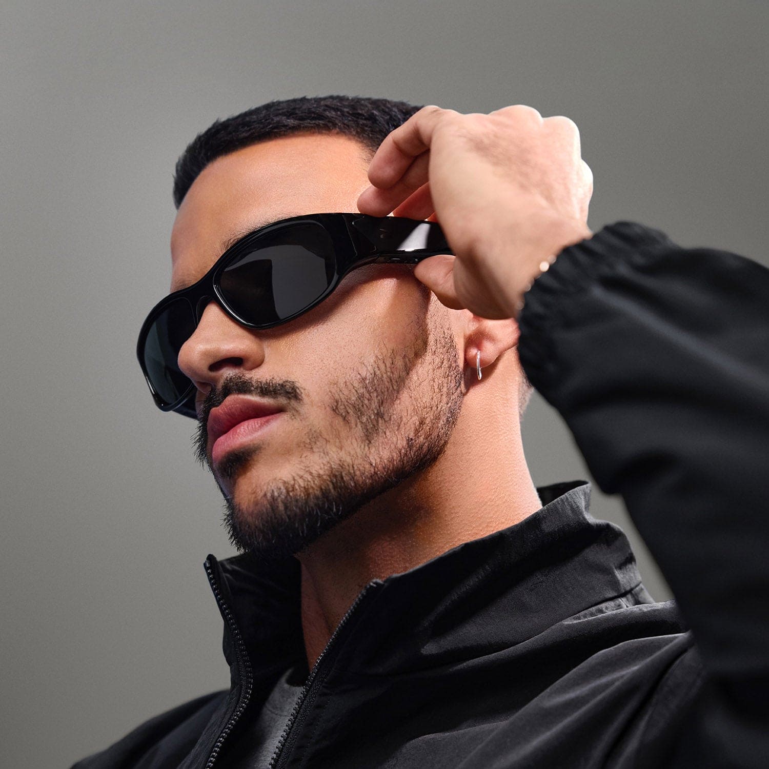 A man with a neatly trimmed beard and mustache, wearing Reebok NITROUS black Bluetooth sport glasses and a black jacket, raises one hand to adjust his glasses against a neutral gray background.