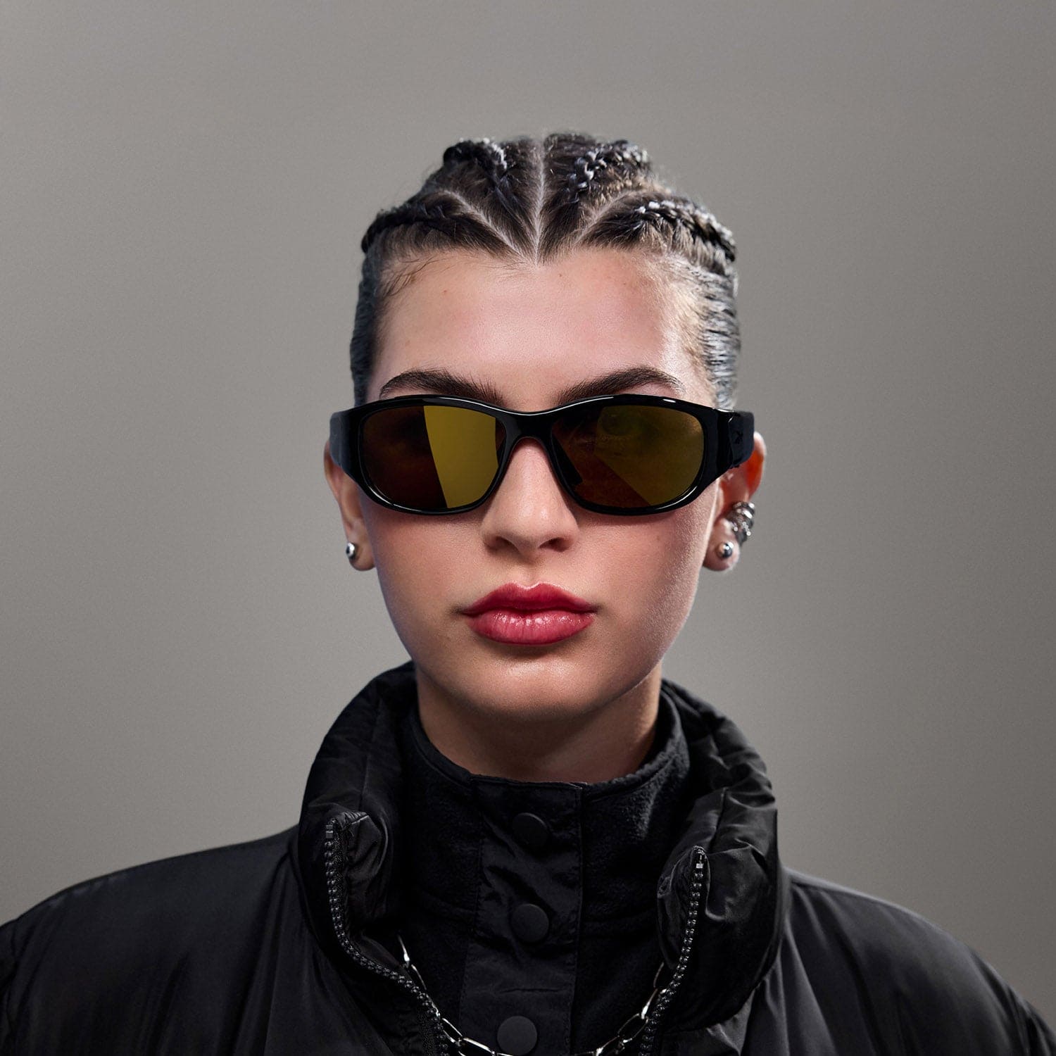 A person with braided hair wears Reebok NITROUS black audio sunglasses with polarized lenses, a black high-collared jacket, and stud earrings while posing against a plain gray background.