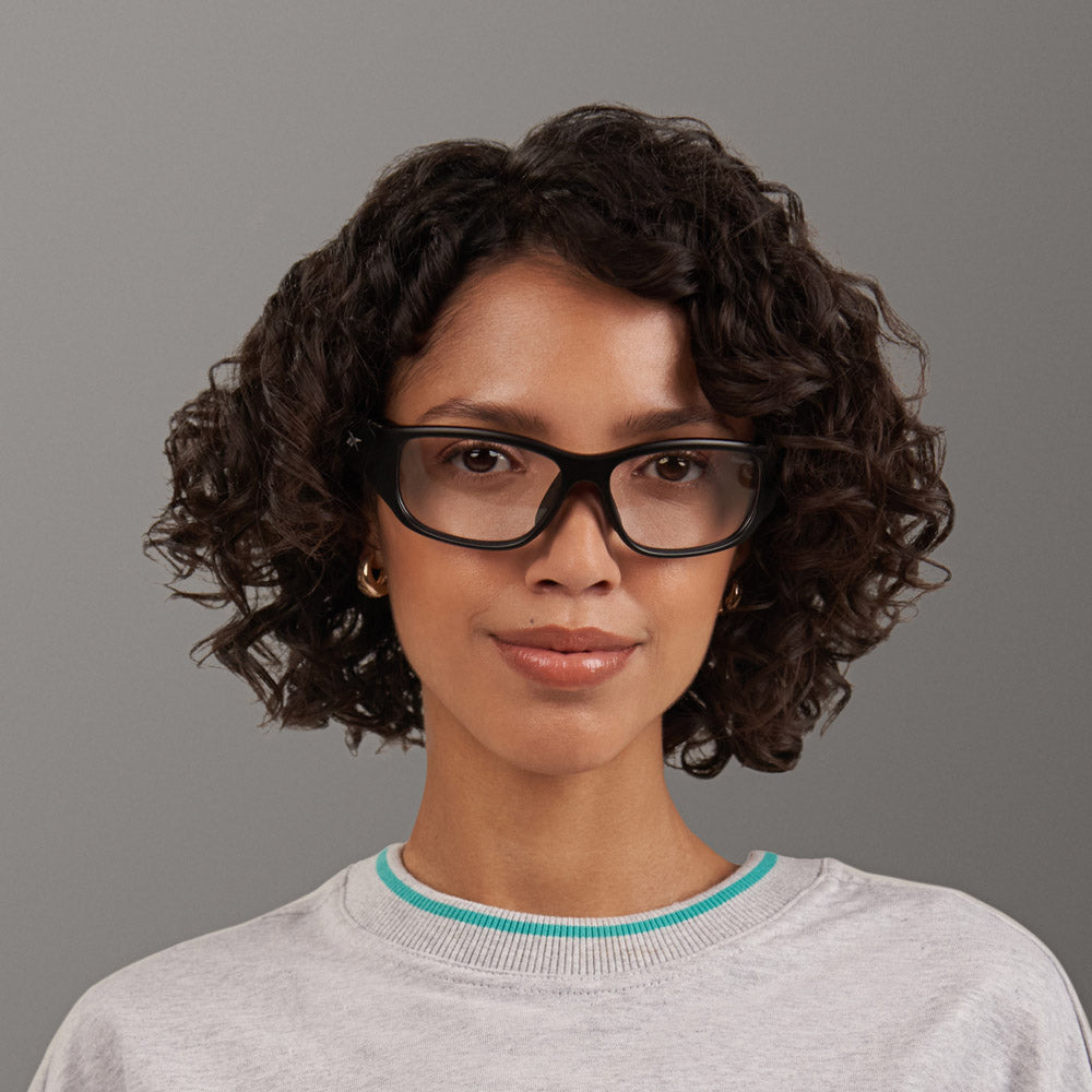 A woman with short curly brown hair wears Reebok Nitrous Shift smart eyewear with adaptive photochromic lenses, gold hoop earrings, and a light gray sweatshirt with turquoise trim, smiling softly against a plain gray background.
