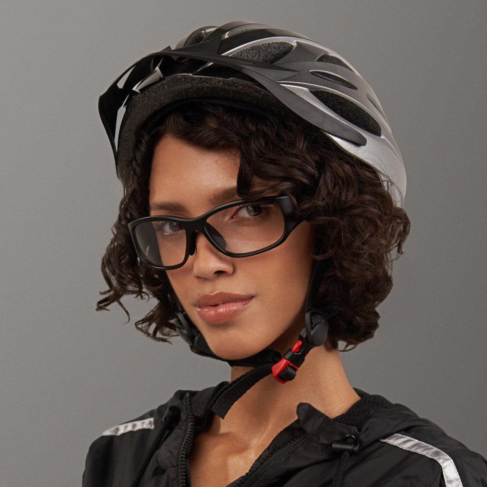 A person with short curly hair wears the Reebok Nitrous Shift black and silver bicycle helmet, smart eyewear with adaptive photochromic lenses, and a black jacket with reflective stripes, confidently facing the camera against a gray background.