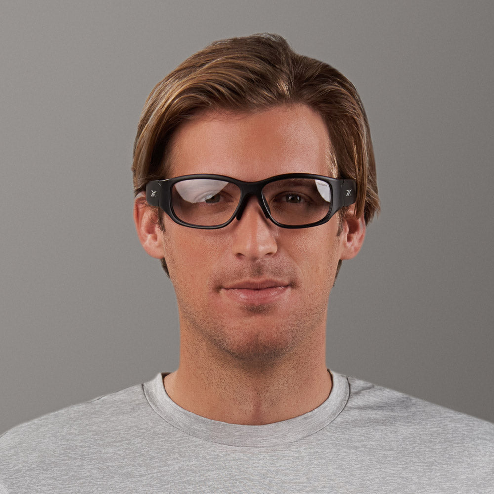 A man with light brown hair wears Reebok Nitrous Shift smart eyewear with adaptive photochromic lenses and a light gray t-shirt, facing the camera against a plain gray background.