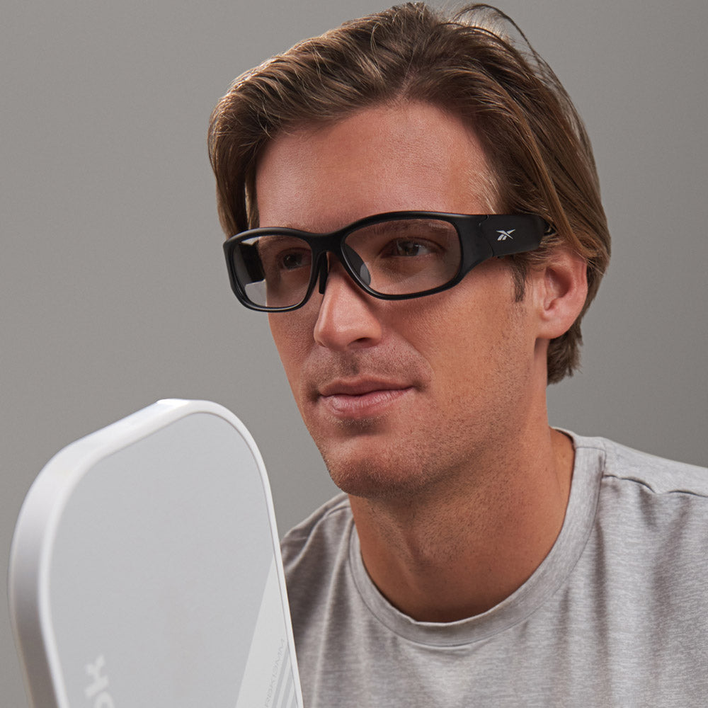 A man wearing Reebok Nitrous Shift smart eyewear with adaptive photochromic lenses and a light gray shirt holds a white pickleball paddle, looking ahead neutrally against a plain gray background.