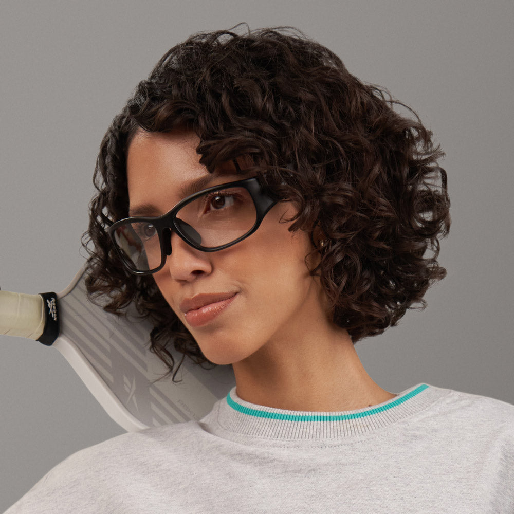 A woman with short, curly dark hair and glasses with adaptive photochromic lenses wears a light gray Reebok Nitrous Shift sweatshirt, holding a tennis racket over her shoulder against a plain gray background.