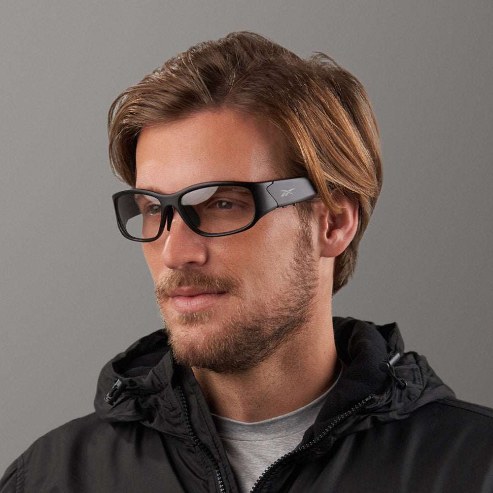 A man with light brown hair and a short beard wears black rectangular smart eyewear and a Reebok Nitrous Shift black zip-up jacket, looking slightly to the side against a plain gray background.