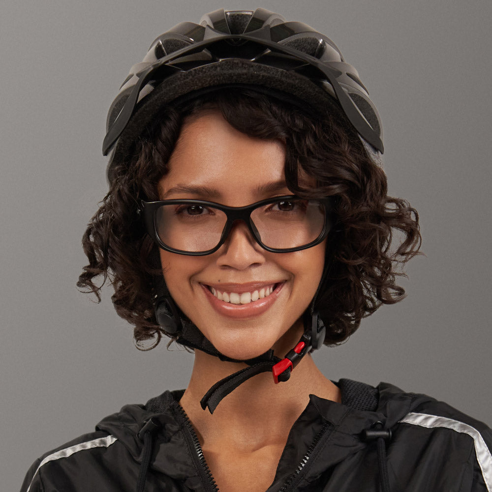 A smiling woman with curly hair wears Reebok Nitrous Shift smart eyewear with adaptive photochromic lenses, a black bike helmet, and a black jacket with white stripes against a plain gray background.