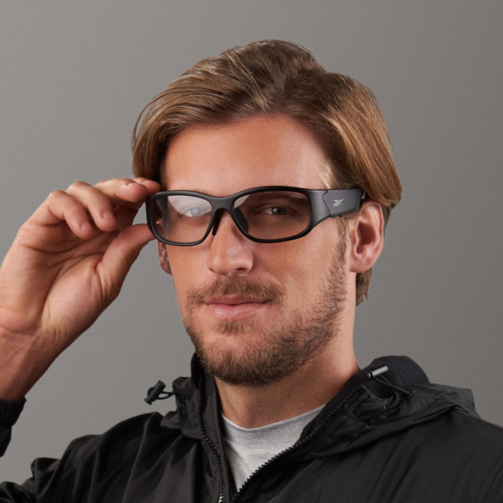 A man with light brown hair and a beard adjusts Reebok Nitrous Shift smart eyewear, featuring adaptive photochromic lenses and black rims, while wearing a black jacket against a plain gray background.