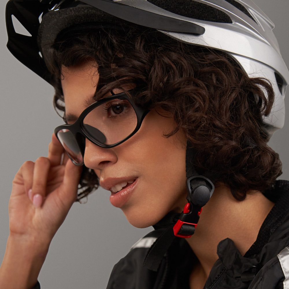 A person with curly hair wearing a white Reebok Nitrous Shift bike helmet and smart eyewear adjusts their glasses while gazing sideways. They're dressed in a black jacket featuring a reflective stripe.