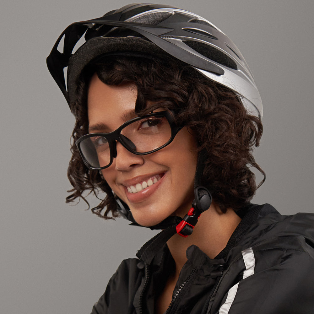 A person with short curly hair and glasses smiles while wearing the Reebok Nitrous Shift bicycle helmet and smart eyewear with adaptive photochromic lenses, plus a black jacket with a white stripe, against a plain gray background.