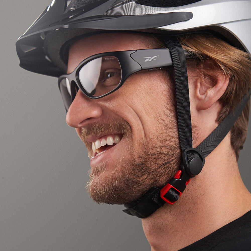 A bearded man smiles while wearing Reebok Nitrous Shift smart eyewear with adaptive photochromic lenses, plus a bike helmet and black chin strap with red buckle, against a plain grey background.