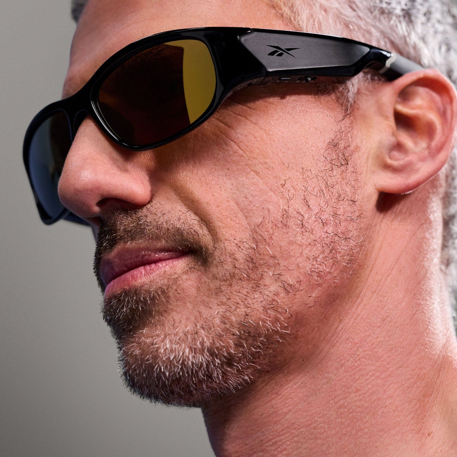 A close-up of a man with short gray hair and stubble wearing Reebok NITROUS large black Bluetooth sport glasses. The neutral background is out of focus.