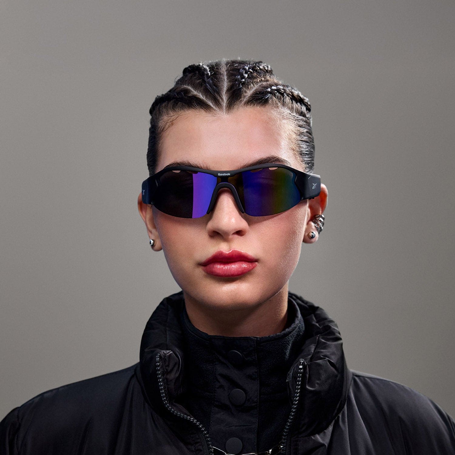 A person with braided hair wears Reebok Octane audio sunglasses with polarized lenses, a black high-collared jacket, and earrings, posing against a plain gray background.