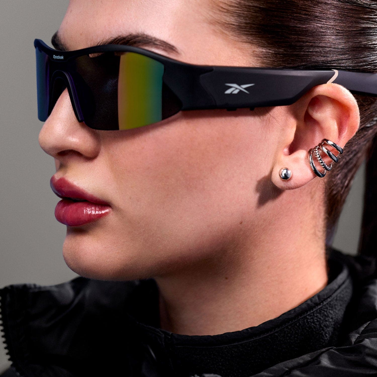 A woman wears Reebok Octane Bluetooth sport sunglasses with mirrored polarized lenses, a pearl stud earring, a multi-ring ear cuff, and a black jacket. Her lips are glossy.