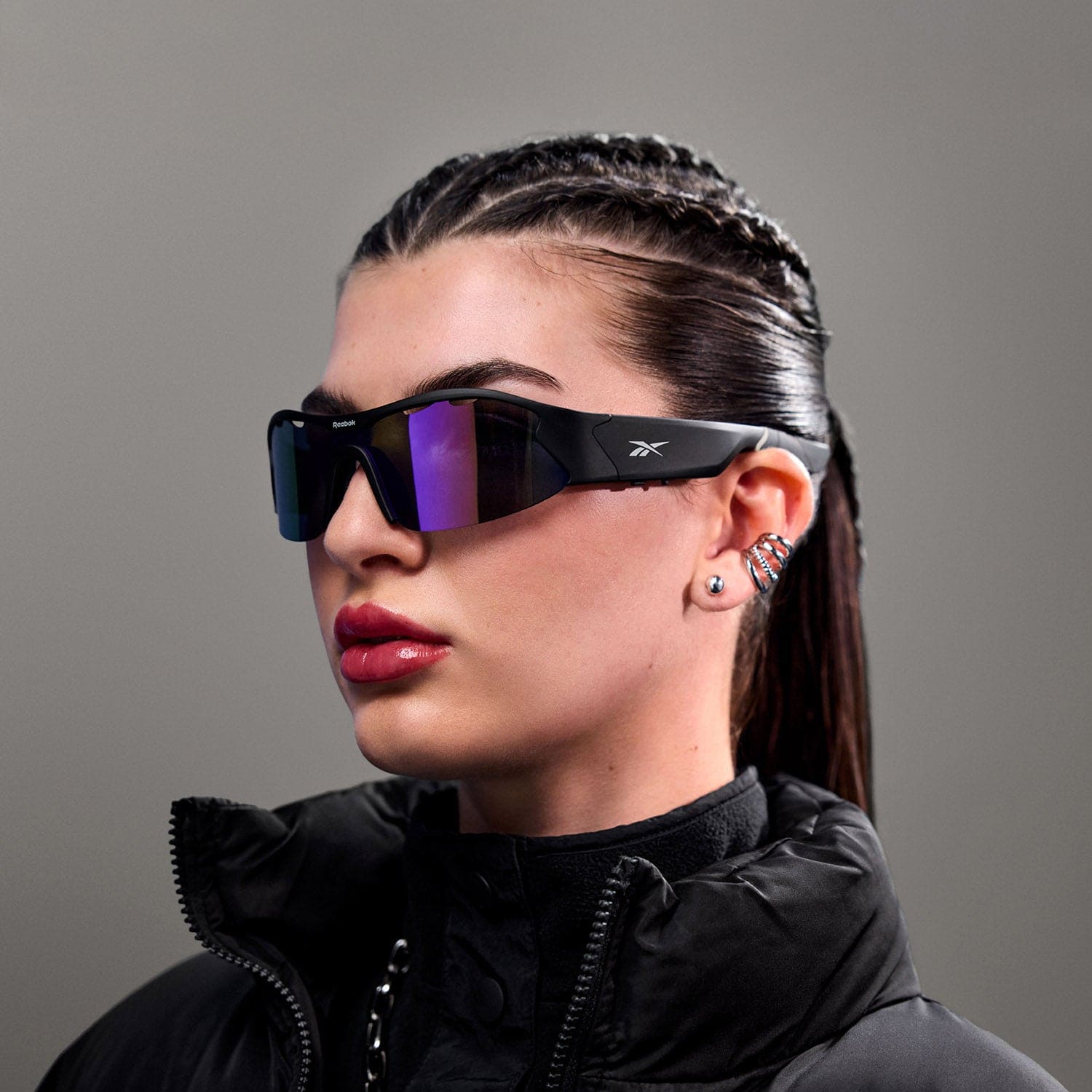 A person with braided hair wears Reebok Octane polarized sports sunglasses, a black puffer jacket, and bold earrings while posing against a plain gray background.
