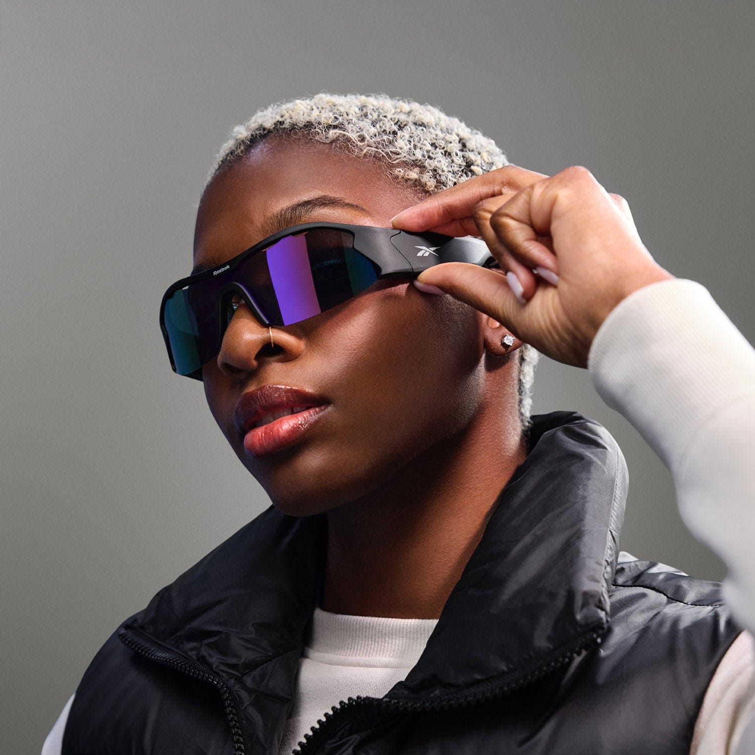 A person with short, light blond hair wears Reebok Octane large, futuristic Bluetooth sport sunglasses with polarized lenses and a black puffer vest, holding the side of the sunglasses with one hand against a neutral background.