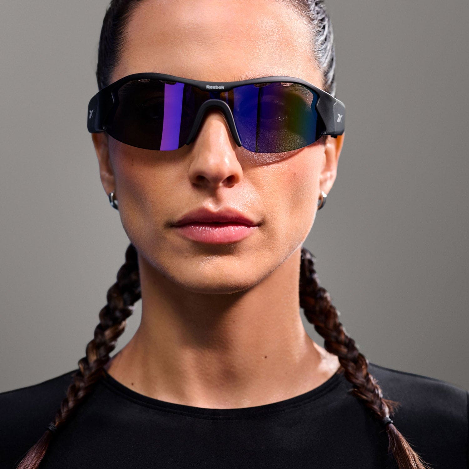 A person with braided hair and a serious expression poses against a gray background, wearing Reebok Octane polarized sunglasses and a black top.