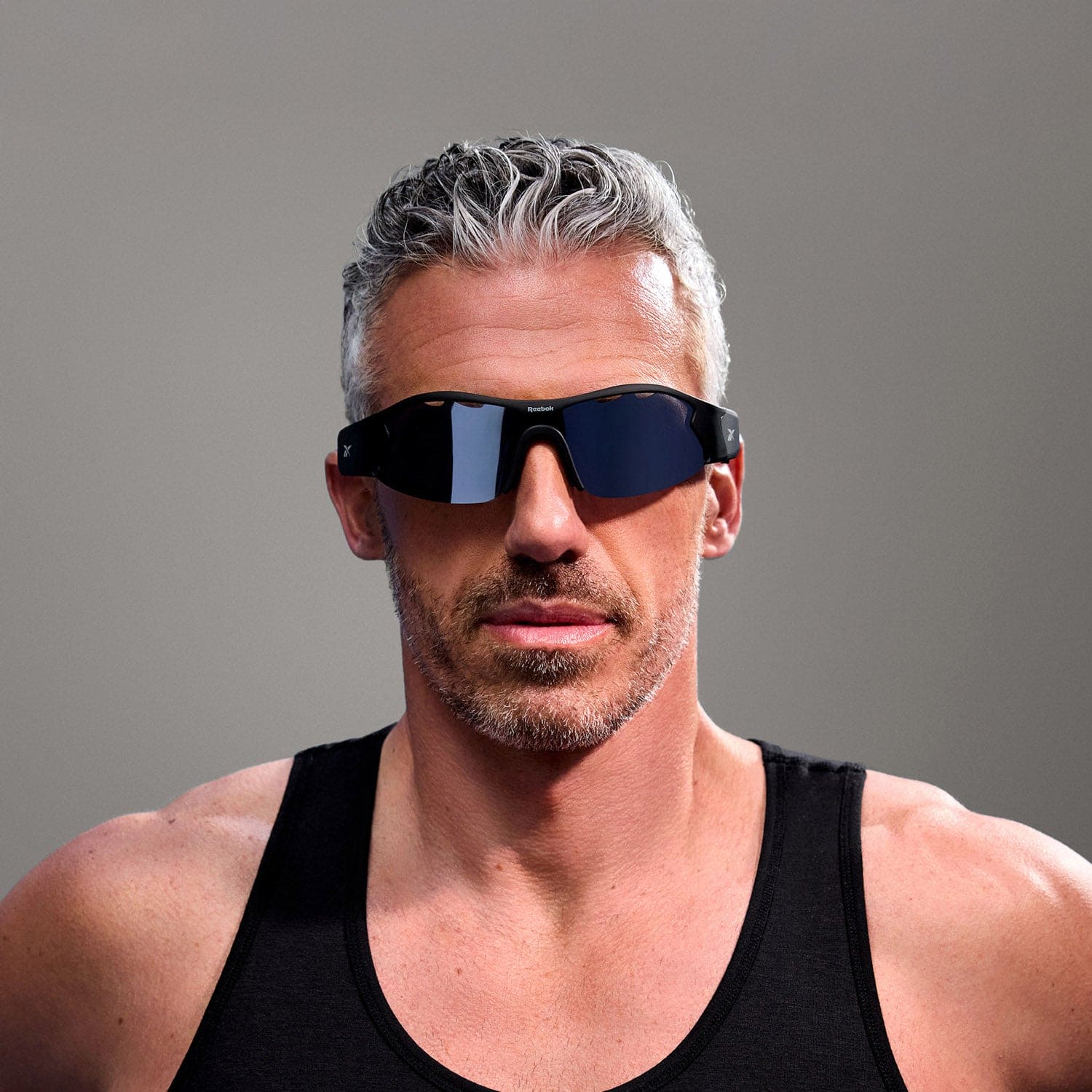 A man with short gray hair and a trimmed beard wears a black Reebok Octane tank top and large polarized audio sunglasses, standing against a plain gray background.