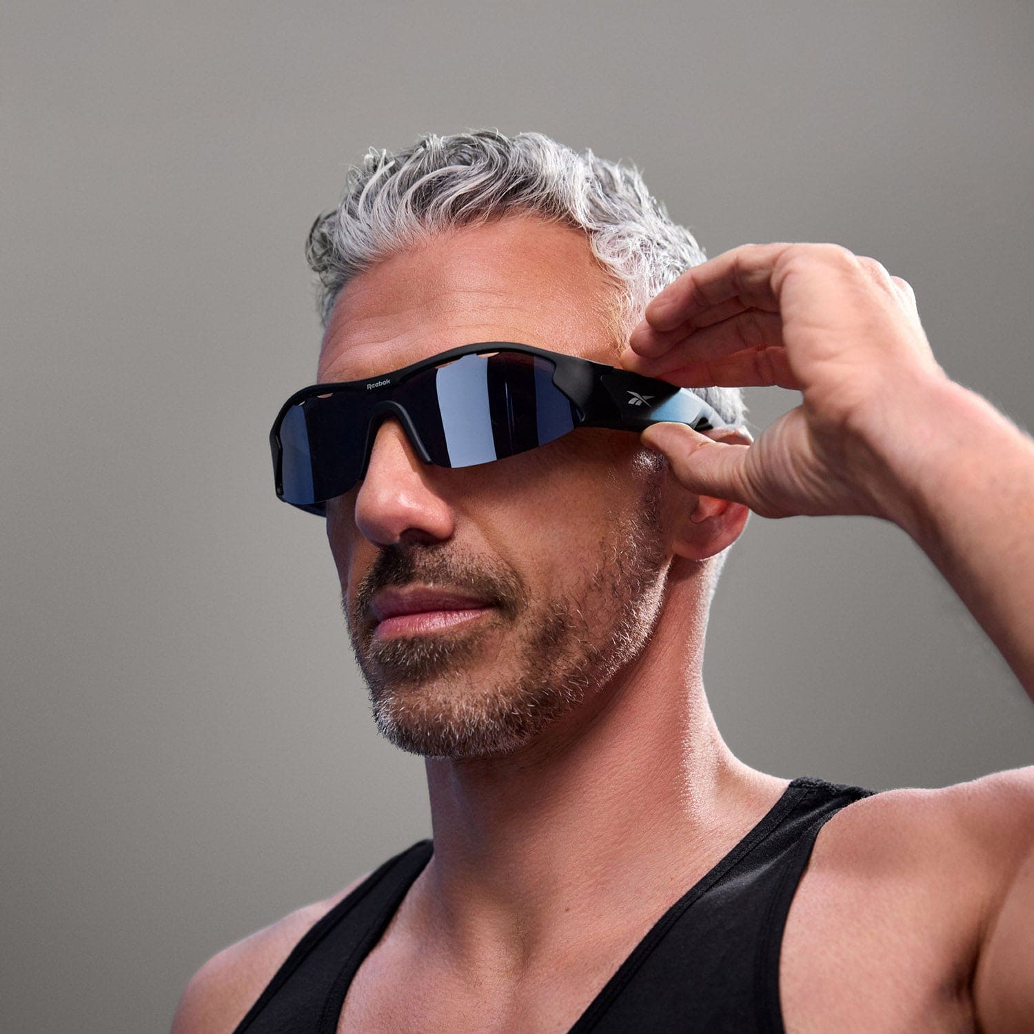 A man with short gray hair and a beard wears Reebok Octane Bluetooth sport sunglasses and a black tank top, adjusting the sunglasses with one hand against a plain gray background.