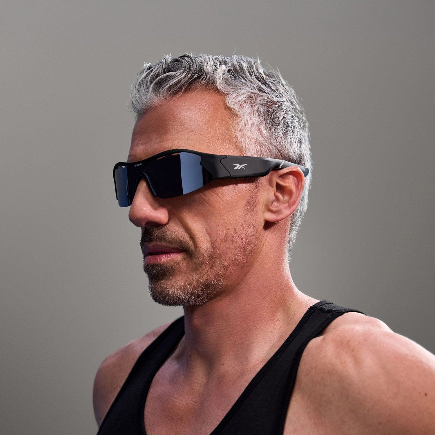 A man with short gray hair and a trimmed beard confidently poses in Reebok Octane black Bluetooth sport sunglasses and a black tank top against a plain gray background.