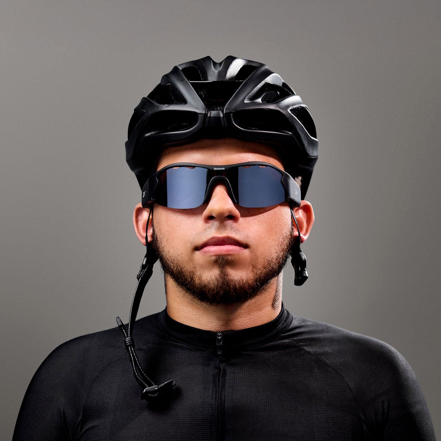 A man wears Reebok's Octane black cycling helmet, Bluetooth sport sunglasses with polarized lenses, and a black cycling jersey, standing against a gray background and looking at the camera.