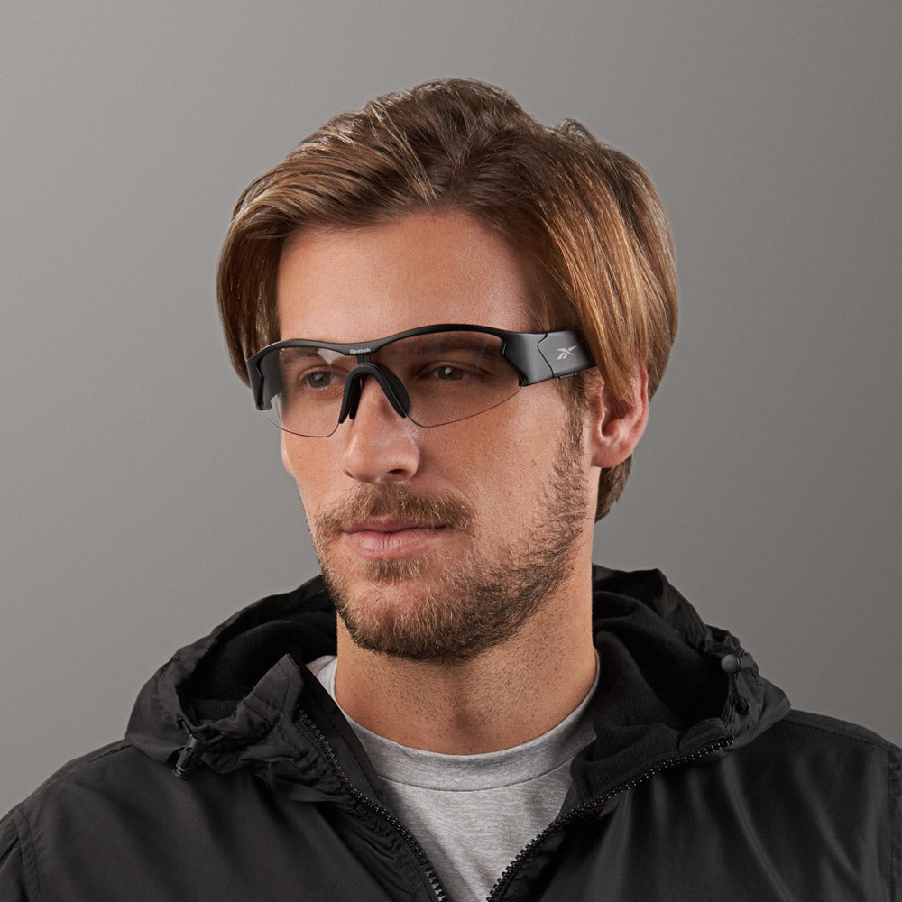 A man with light brown hair and a beard wears Reebok Octane Shift smart eyewear with tinted photochromic lenses and a black jacket, posing against a plain gray background.