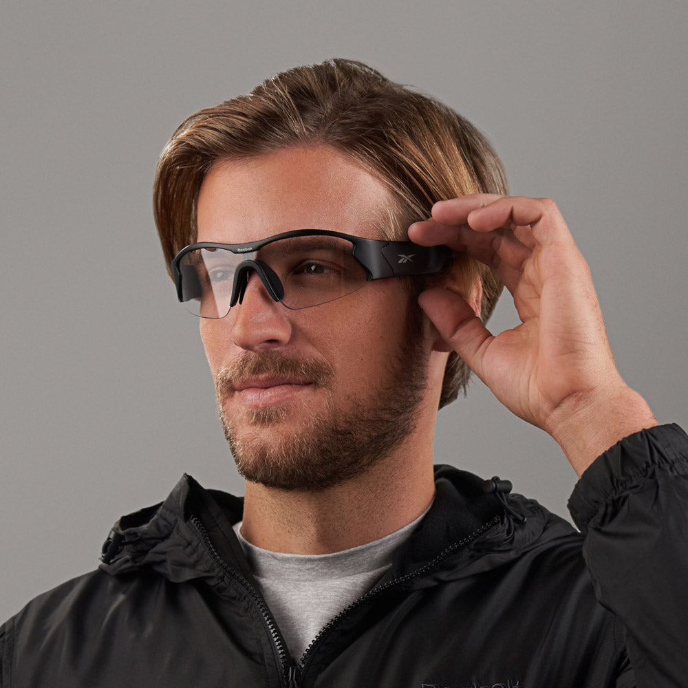 A light-brown-haired man with a beard wears Reebok Octane Shift sporty black smart eyewear and a black jacket, touching the glasses' side with his right hand against a plain gray background.