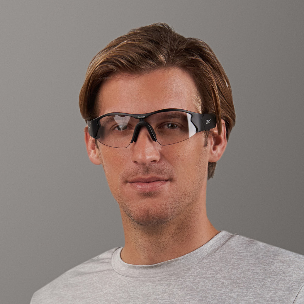 A man with light brown hair wears a light gray t-shirt and Reebok Octane Shift black smart eyewear with photochromic lenses, standing before a plain gray background and looking straight at the camera with a neutral expression.