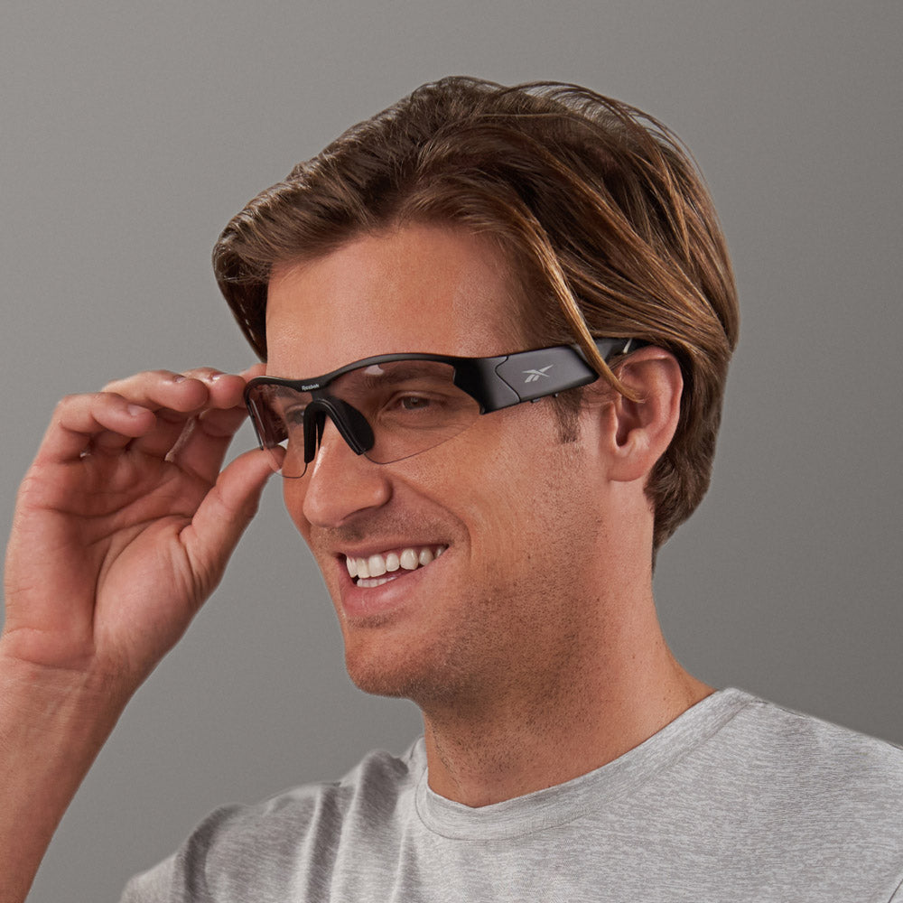 A smiling person with light brown hair wears Reebok Octane Shift black smart eyewear and a light grey t-shirt, adjusting the glasses with their right hand against a plain grey background.