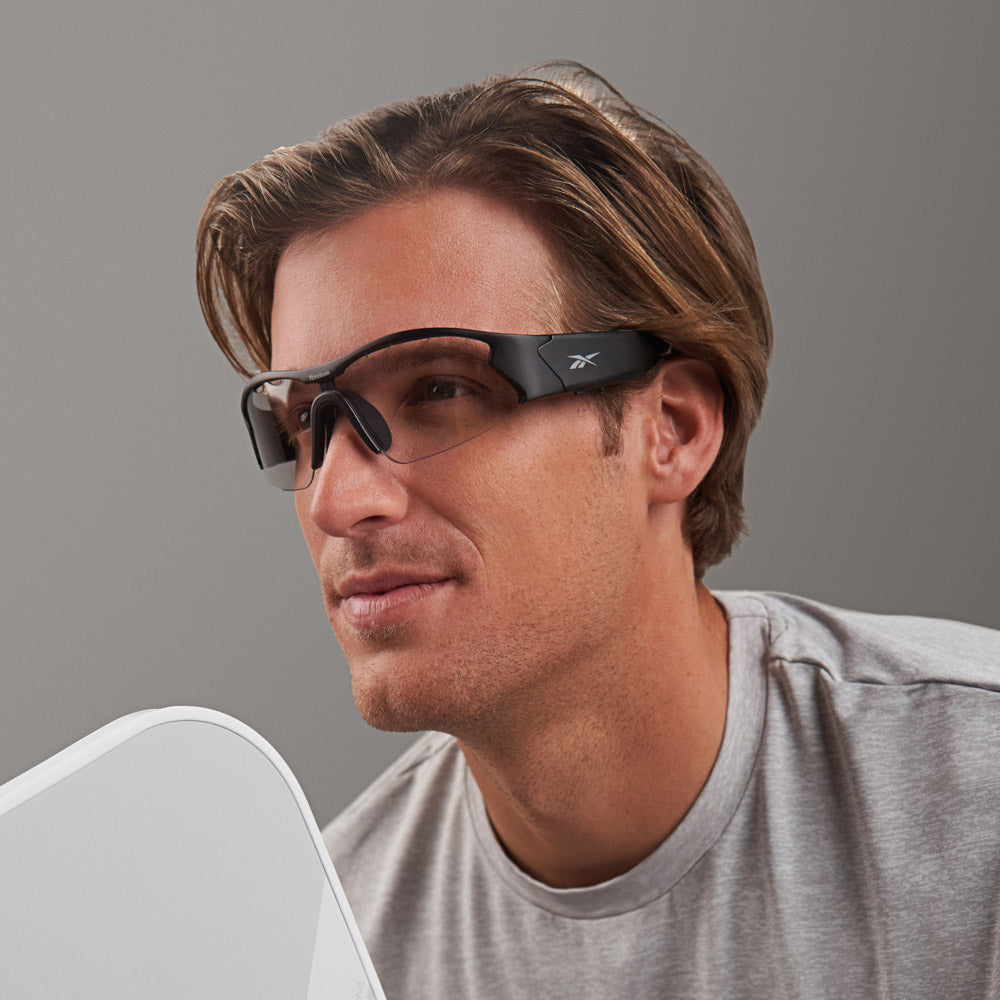 A man with light brown hair wears Reebok Octane Shift black smart eyewear with photochromic lenses and a gray t-shirt, looking off to the side against a plain gray background.