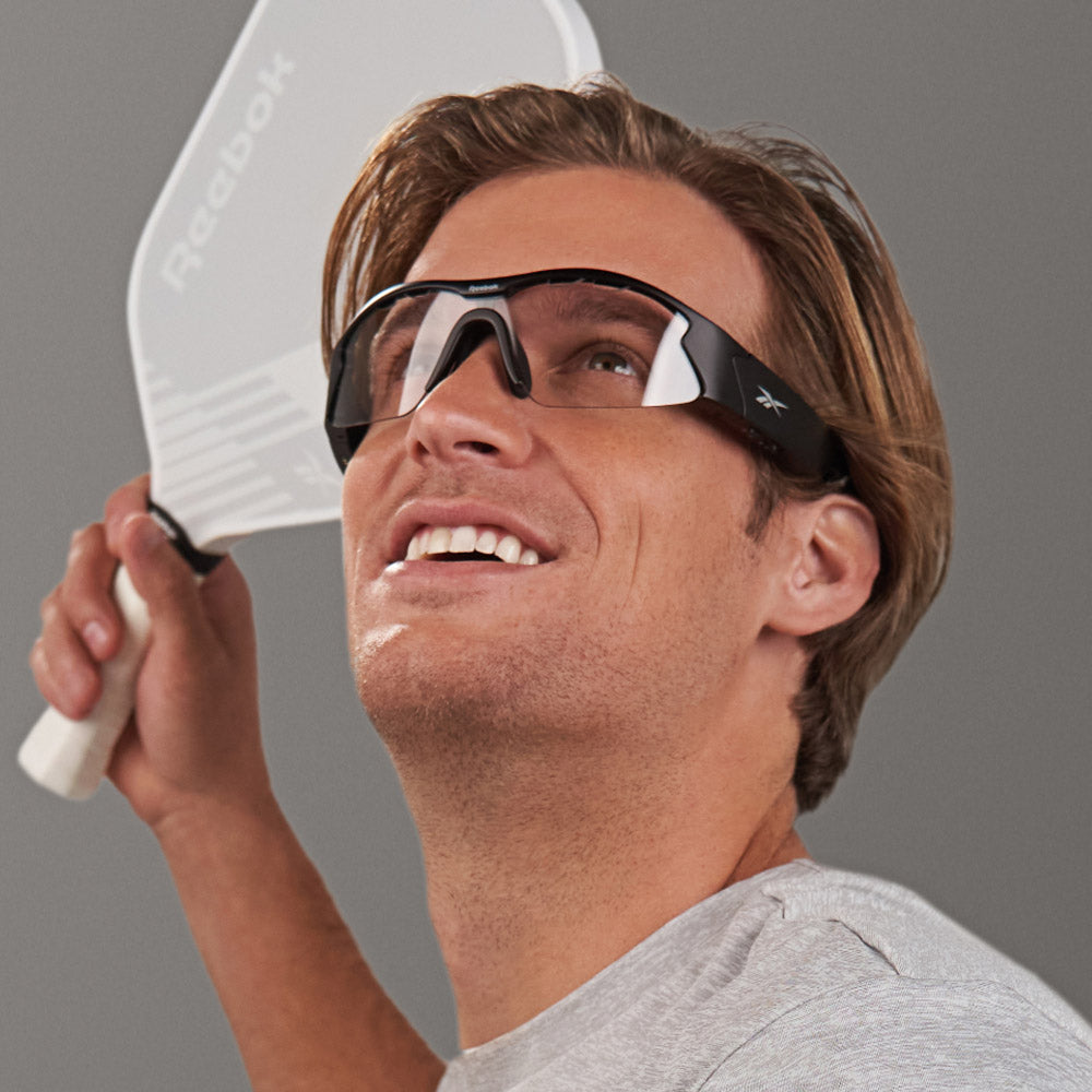 A smiling man in a light gray shirt and Octane Shift smart eyewear with photochromic lenses holds a white Reebok paddle, poised to play, against a plain gray background.