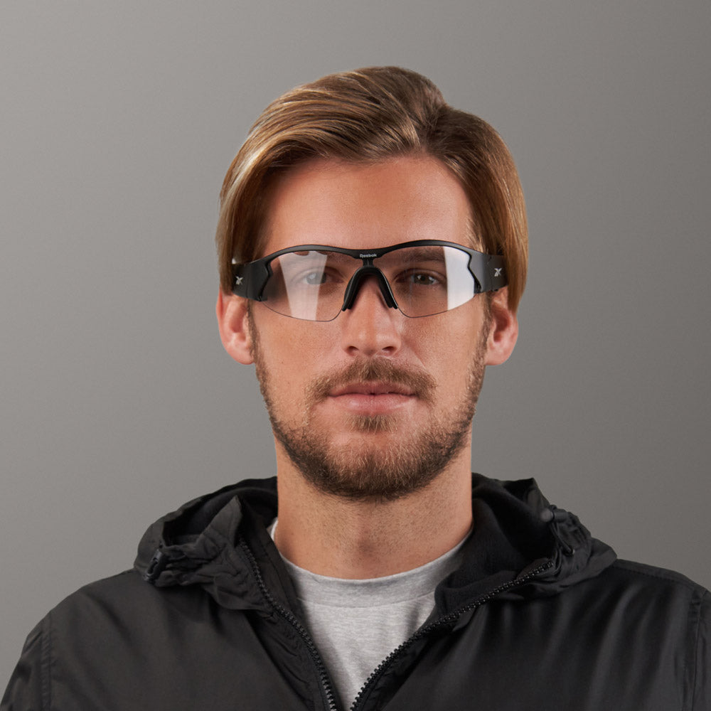 A man with light brown hair and a beard wears Reebok Octane Shift smart eyewear with photochromic lenses, along with a gray shirt and black jacket, standing against a plain gray background.