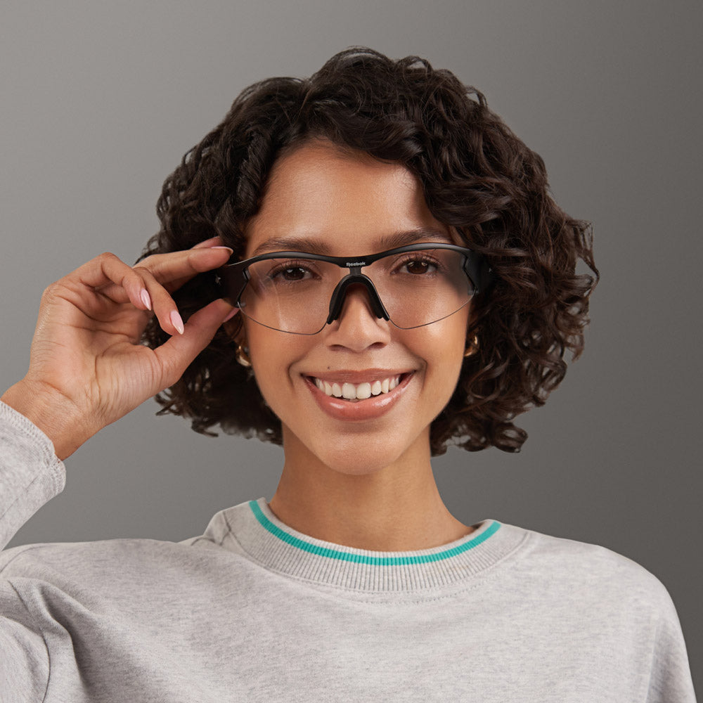 A smiling person with short curly hair wears a light gray sweatshirt and Reebok’s Octane Shift smart eyewear, holding the frames with one hand, against a plain gray background.