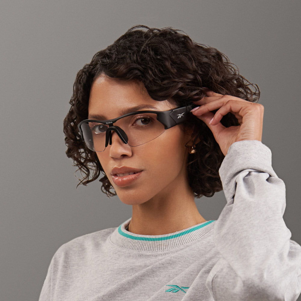 A woman with short curly hair wears sporty black Reebok Octane Shift smart eyewear and a grey Reebok sweatshirt, posing against a plain grey background.