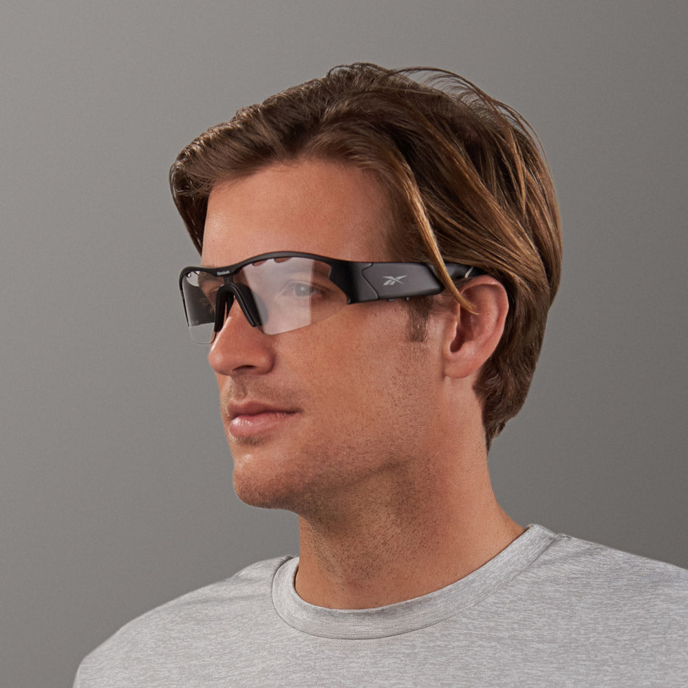 A man with light brown hair, in a light grey shirt and large, dark Reebok Octane Shift photochromic lenses, poses against a plain grey background.