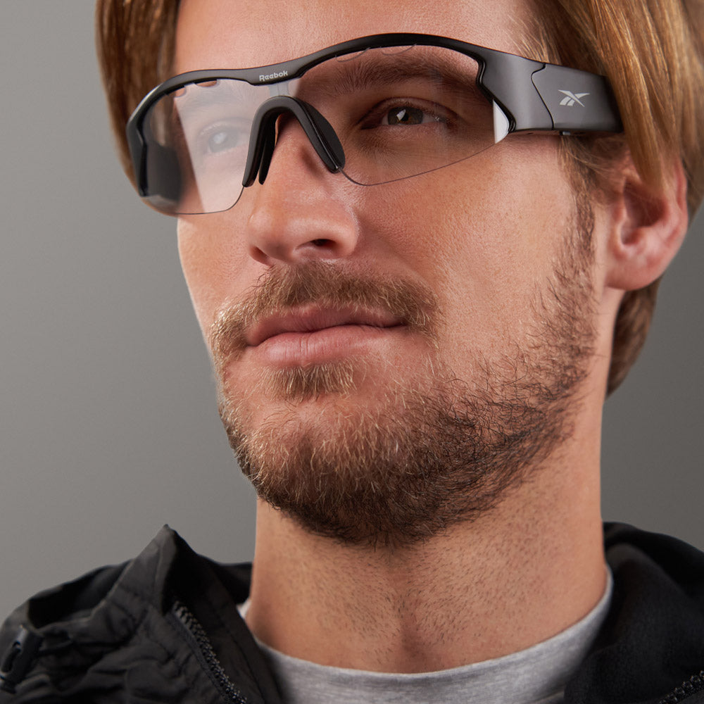 A man with light brown hair and a beard wears black Reebok Octane Shift smart eyewear sports sunglasses and a black jacket, looking slightly to the side against a plain gray background.