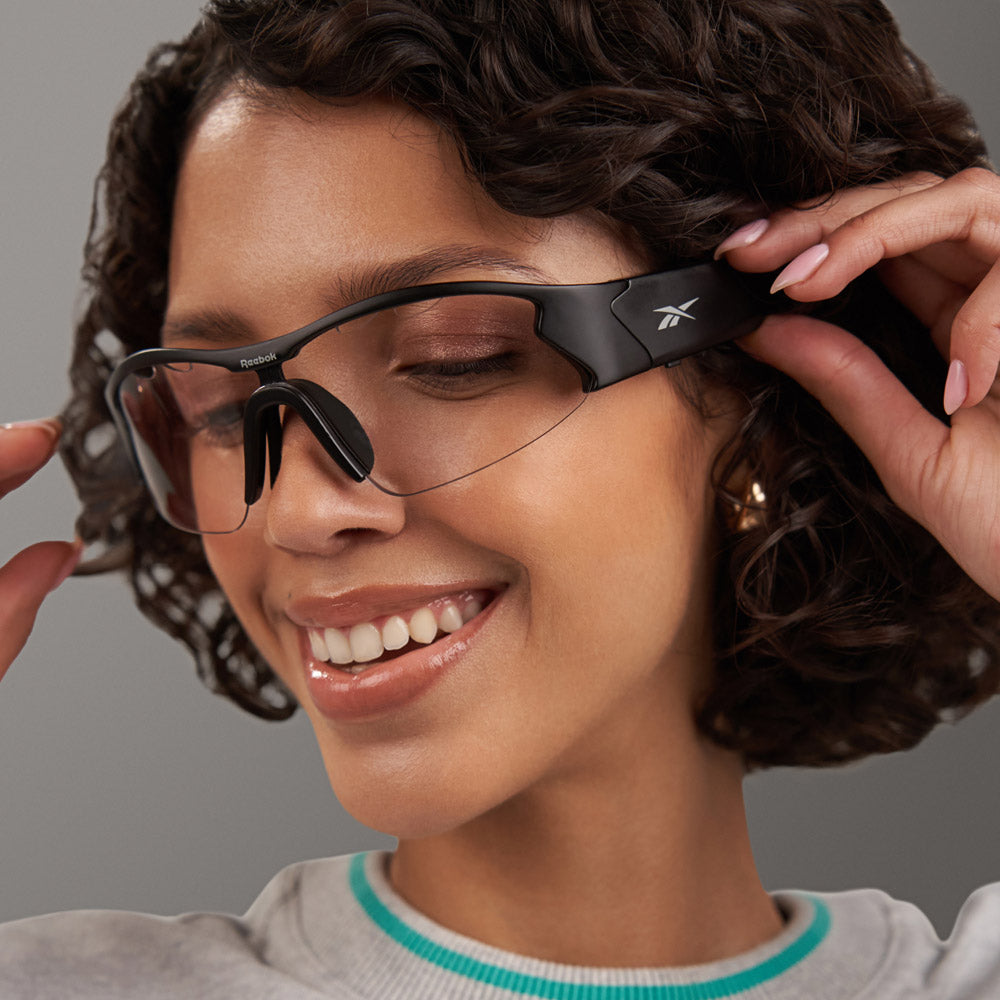 A woman with curly hair smiles while adjusting Reebok Octane Shift smart eyewear sunglasses. She wears hoop earrings and a light top, looking happy and relaxed against a neutral background.